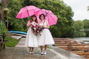 Lesbian brides in wedding dresses standing under pink umbrellas