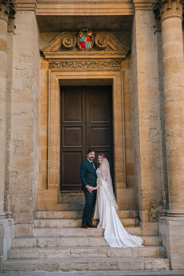 Bride and groom standing outside the Sheldonian Theatre in Oxford, framed by golden stone and Gothic arches.