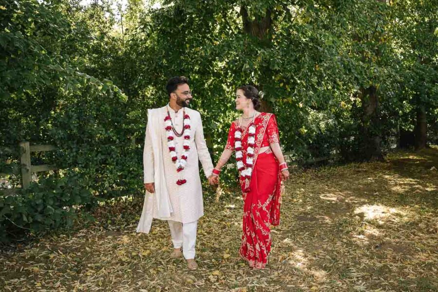 Elegant Indian wedding couple holding hands in a lush garden, traditional attire, floral garlands, romantic outdoor ceremony, Rachel Movitz Weddings.